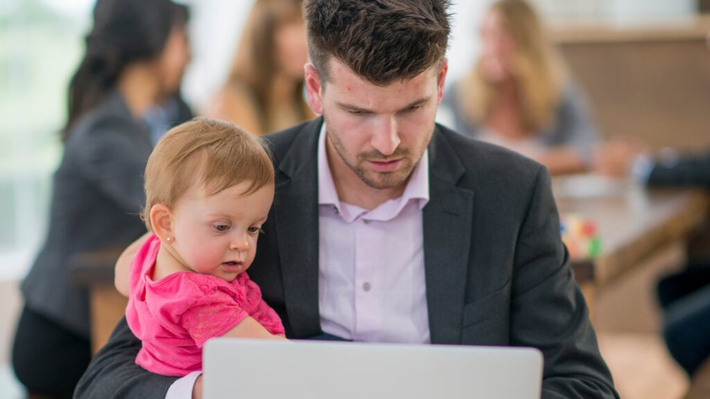 A Working Parent on Laptop while his holding baby girl in one arm