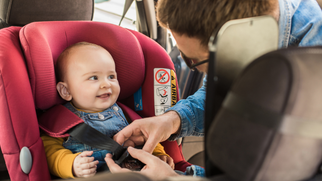 A father strapping his baby in a red coloured rear facing car seat.